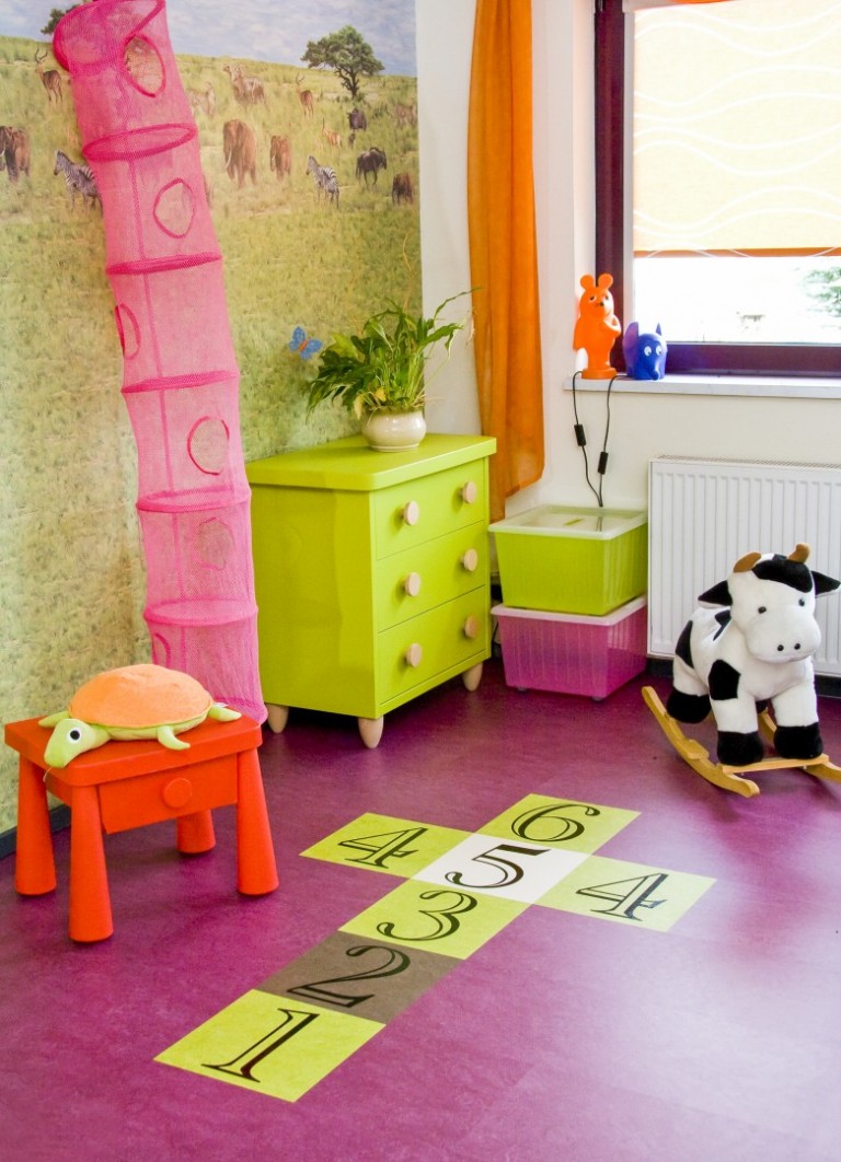 Farbenfrohes Kinderzimmer mit grüner Kommode, rosa Turm, schaukelnder Kuh, Spielzeugschildkröte auf rotem Tisch und Hüpfkästchen auf dem lila Laminatboden. -Bodenstyle Schuster - Industriestraße 2 - 66802 Überherrn - +49 (0)6836-7789955
