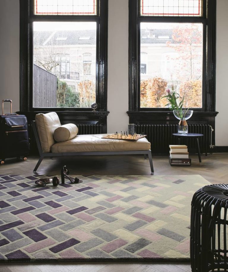 Modernes Wohnzimmer mit beiger Chaiselongue, Schachbrett, Gepäck, gemustertem Teppich auf stilvollem Parkettboden, rundem Tisch mit Büchern und Vase und großen Fenstern, die den Blick auf Bäume und städtische Gebäude freigeben. -Bodenstyle Schuster - Industriestraße 2 - 66802 Überherrn - +49 (0)6836-7789955