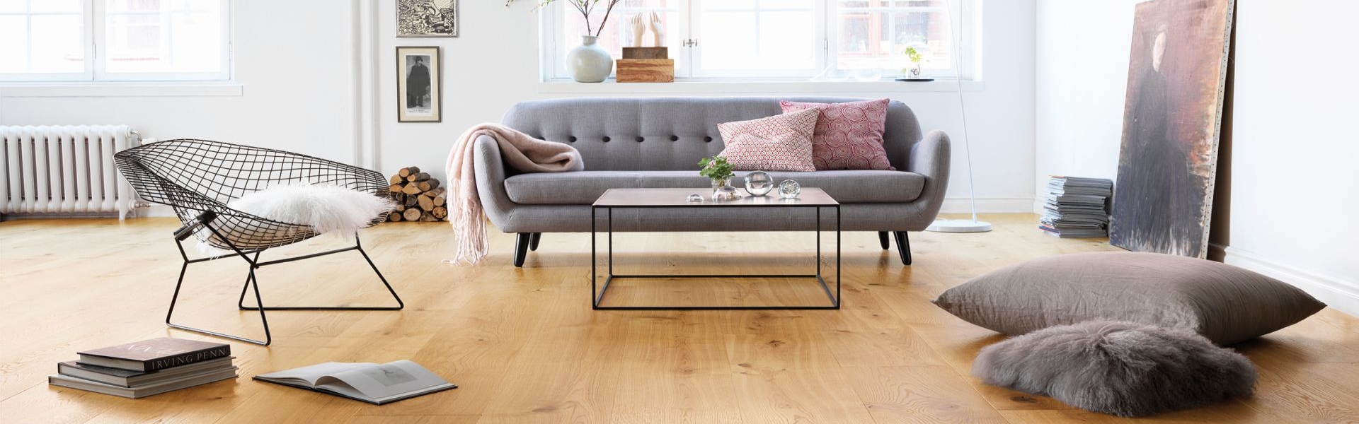 Modernes Wohnzimmer mit grauem Sofa, Drahtstuhl, Couchtisch, Bodenkissen und Büchern auf schönem Parkettboden. Große Fenster sorgen für natürliches Licht. -Bodenstyle Schuster - Industriestraße 2 - 66802 Überherrn - +49 (0)6836-7789955