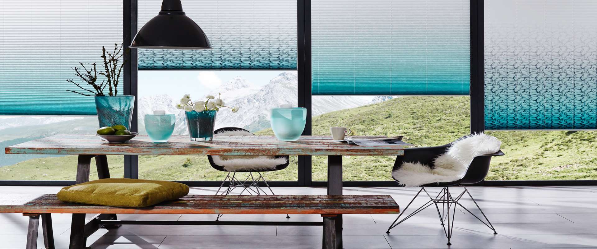 Ein rustikaler Holztisch mit Glasvasen, eine Bank und Stühle mit Fellüberwürfen stehen auf stilvollen Bodenbelägen vor großen Fenstern mit blauen Jalousien, die den Blick auf eine Berglandschaft freigeben. -Bodenstyle Schuster - Industriestraße 2 - 66802 Überherrn - +49 (0)6836-7789955