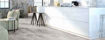 Moderne Küche mit weißen Schränken, einer langen Arbeitsplatte, Barhockern und einem offenen, industriell anmutenden Interieur mit großen Fenstern und Betonboden - perfekt für einen Bodenleger, der stilvolles Laminat oder Parkett verlegt. -Bodenstyle Schuster - Industriestraße 2 - 66802 Überherrn - +49 (0)6836-7789955