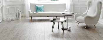 Ein modernes Wohnzimmer mit weißen Wänden, einem weißen Sofa, einem weißen Sessel, zwei runden Tischen und stilvollem Laminatfußboden. Ein blau gemustertes Kissen liegt auf dem Sofa. -Bodenstyle Schuster - Industriestraße 2 - 66802 Überherrn - +49 (0)6836-7789955
