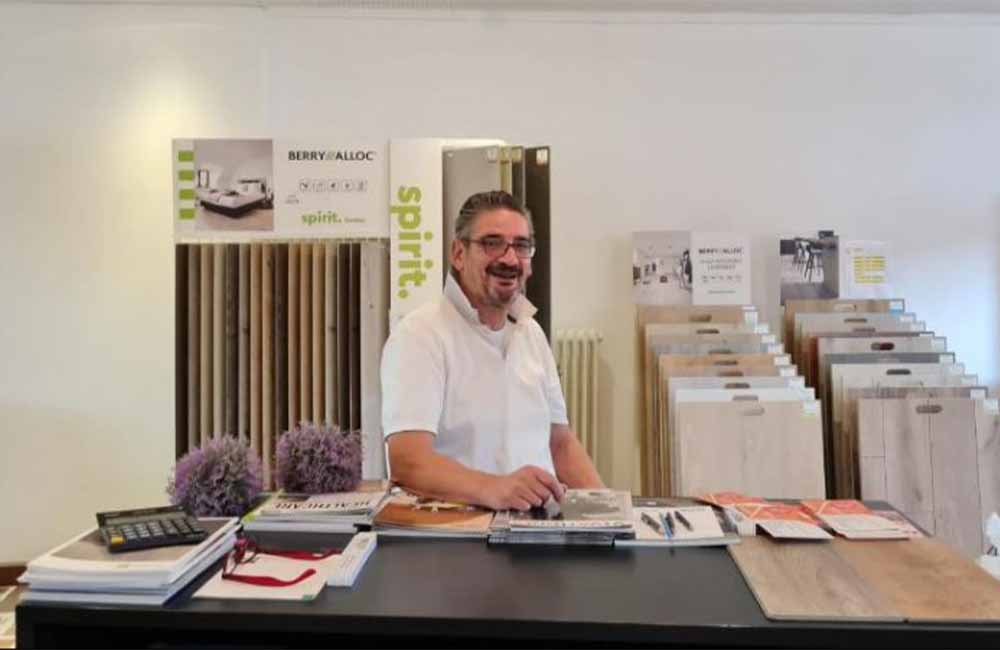 Ein Mann in einem weißen Hemd steht hinter einer Theke mit Teppichboden- und Laminatmustern, Broschüren und Büromaterial in einem Ausstellungsraum für Bodenbeläge. -Bodenstyle Schuster - Industriestraße 2 - 66802 Überherrn - +49 (0)6836-7789955