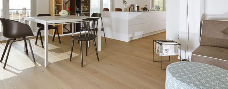 Moderner Essbereich mit weißem Tisch, schwarzen Stühlen und hellem Laminatboden, der den Blick auf eine helle, minimalistische Küche im Hintergrund freigibt. -Bodenstyle Schuster - Industriestraße 2 - 66802 Überherrn - +49 (0)6836-7789955