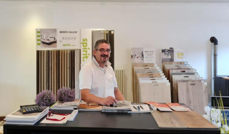 Ein Mann mit Brille und weißem Polohemd steht hinter einem Tresen in einem Ausstellungsraum für Bodenbeläge, umgeben von Broschüren und Holzmustern, die Bodenbeläge wie Laminat und Parkett präsentieren. -Bodenstyle Schuster - Industriestraße 2 - 66802 Überherrn - +49 (0)6836-7789955