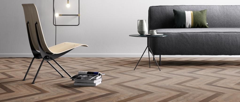 Minimalistisches Wohnzimmer mit einem Parkettboden im Chevron-Muster, einem modernen Sessel, einem kleinen runden Tisch mit Krug und Glas, Büchern und einem grauen Sofa mit Kissen - ein einladender Raum, der durch fachmännisches Bodenleger-Handwerk gestaltet wurde. -Bodenstyle Schuster - Industriestraße 2 - 66802 Überherrn - +49 (0)6836-7789955