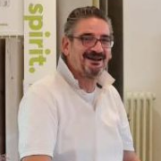 Ein Mann mit Brille und weißem Hemd steht drinnen und lächelt, im Hintergrund ist ein Schild mit der Aufschrift "spirit" zu sehen - vielleicht in einem Ausstellungsraum für Bodenbeläge, in dem ein geschickter Bodenleger stilvolle Vinylbeläge anbringt. -Bodenstyle Schuster - Industriestraße 2 - 66802 Überherrn - +49 (0)6836-7789955