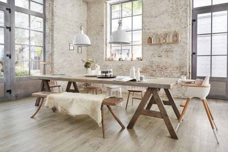 Das rustikale Esszimmer verfügt über einen langen Holztisch mit Bänken, moderne Stühle, Pendelleuchten, große Fenster und einen stilvollen Vinylbelag, der von einem erfahrenen Bodenleger verlegt wurde und für zusätzlichen Charme und Haltbarkeit sorgt. -Bodenstyle Schuster - Industriestraße 2 - 66802 Überherrn - +49 (0)6836-7789955
