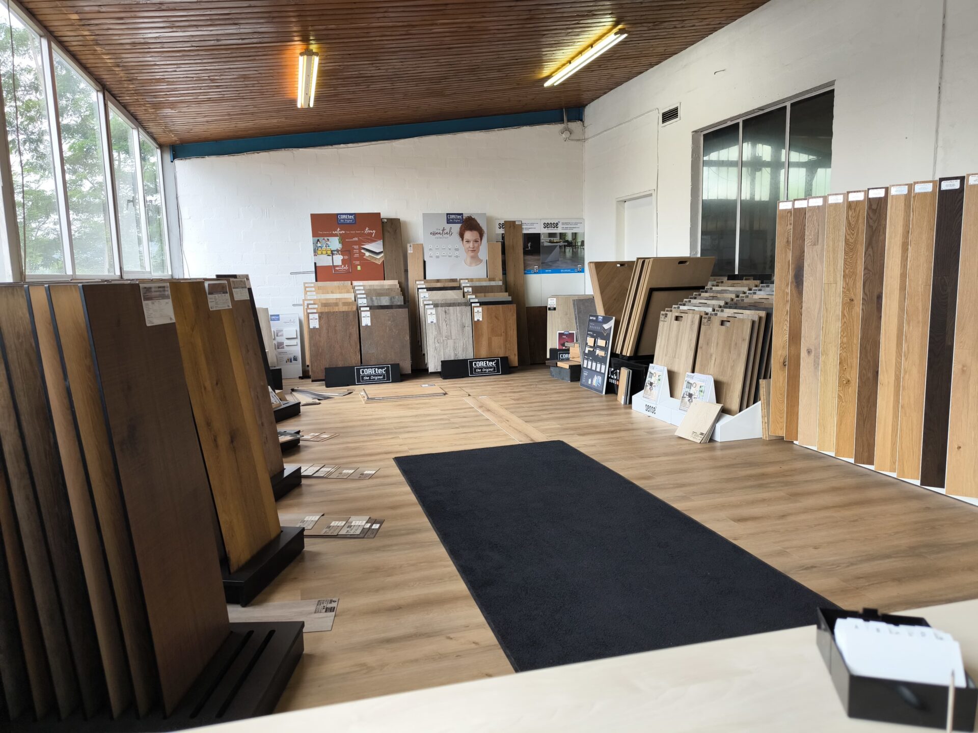 Ausstellungsraum mit verschiedenen Parkett- und Laminat-Bodenmustern auf Ausstellungsregalen, großen Fenstern, holzgetäfelter Decke und Werbepostern - die perfekte Inspiration für Ihr nächstes Bodenleger-Projekt. -Bodenstyle Schuster - Industriestraße 2 - 66802 Überherrn - +49 (0)6836-7789955